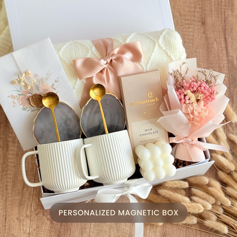 May include: A gift box with a white lid, containing two white ribbed mugs with gold spoons, coasters, a small bouquet of pink flowers, chocolate, and a cream-colored blanket tied with a pink bow. The box is labeled "PERSONALIZED MAGNETIC BOX".