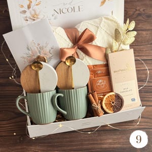 May include: A gift box with a cream-colored sweater tied with a peach ribbon, two green mugs, and two coasters with gold spoons. The box also contains a chocolate bar, cinnamon sticks, and a dried orange slice.