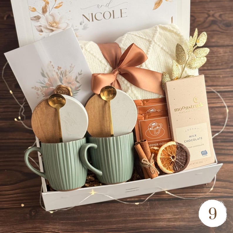 May include: A gift box with a cream-colored sweater tied with a peach ribbon, two green mugs, and two coasters with gold spoons. The box also contains a chocolate bar, cinnamon sticks, and a dried orange slice.