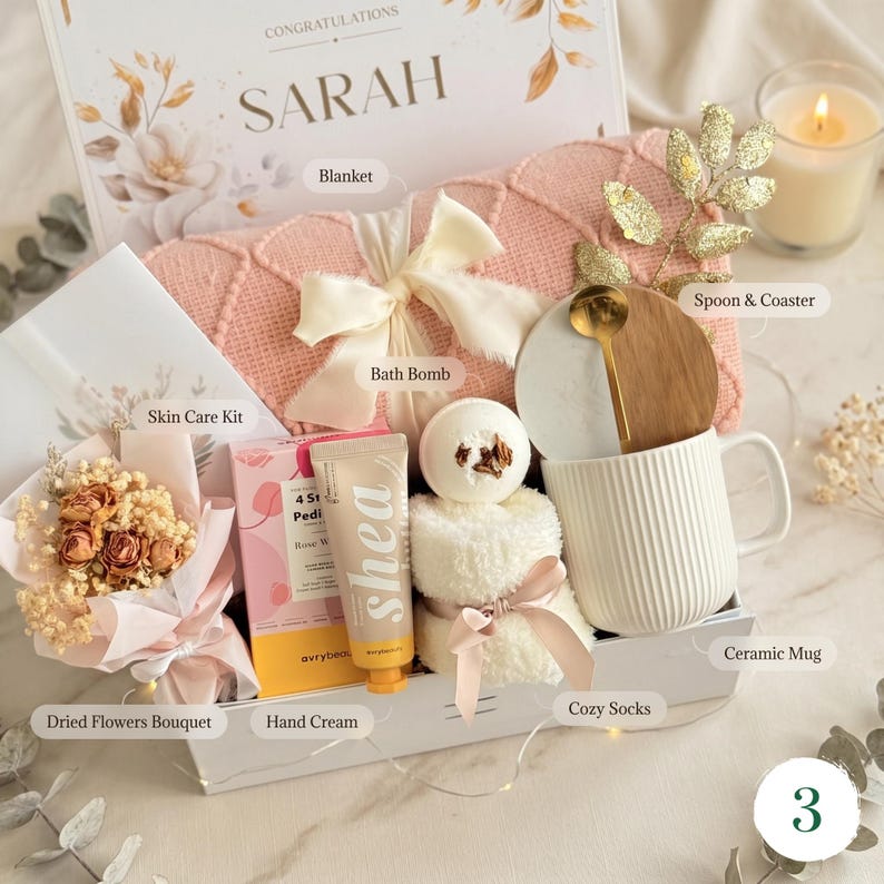 May include: A gift box containing a pink blanket, bath bomb, skincare kit, dried flowers, hand cream, cosy socks, a ceramic mug, and a spoon with a coaster. The box is decorated with a bow and the name "SARAH".