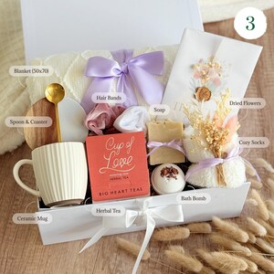 May include: A white gift box filled with self-care items, including a ceramic mug, herbal tea, soap, bath bomb, hair bands, a blanket, cozy socks, and dried flowers. The box is tied with a purple ribbon and has the words "Cup of Love" on the side.