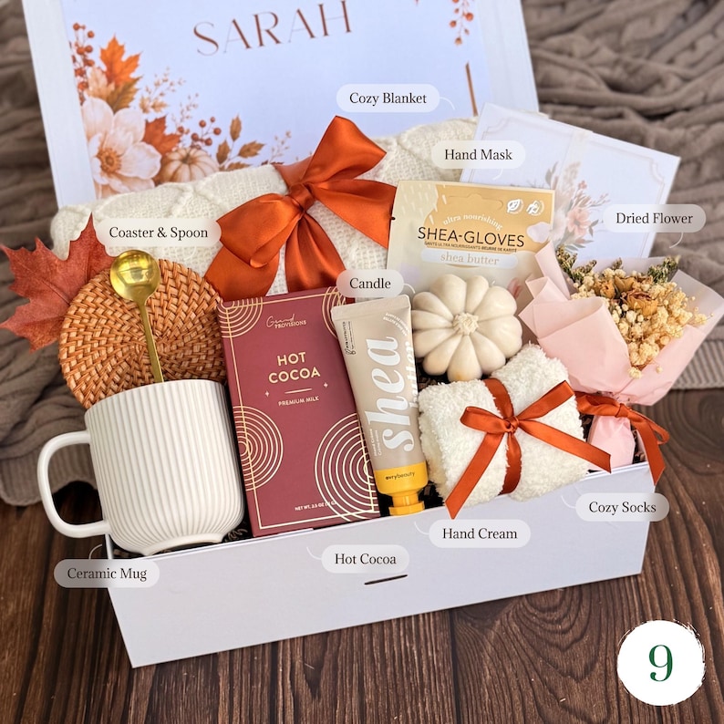May include: A gift box containing a white ribbed ceramic mug, a gold spoon, a woven coaster, a box of hot cocoa, a tube of shea hand cream, a shea glove hand mask, a pumpkin-shaped candle, white fuzzy socks with a ribbon, and dried flowers.