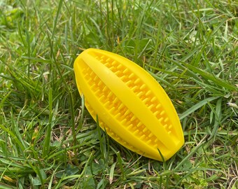 May include: A bright yellow dog toy shaped like a corn cob, lying on a bed of green grass. The toy has a textured surface with rows of small, raised nubs. The overall design is simple and functional, suggesting durability and playfulness.