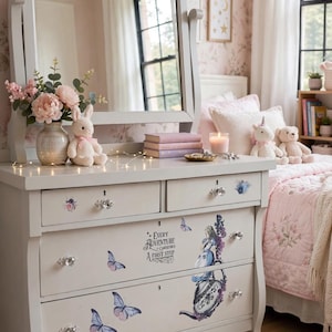 May include: A light gray wooden dresser with four drawers, featuring crystal-style knobs and decorative decals of butterflies, a teapot, and a character. The text reads: Every adventure requires a first step. The dresser includes a matching mirror.
