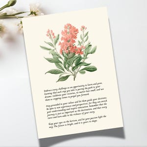 May include: A rectangular card with a cream background featuring a botanical illustration of pink flowers with green leaves at the top. Below the illustration, black text provides an inspirational message. The card is positioned next to two pens.