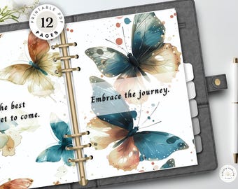 May include: A planner with ring binding featuring watercolor butterfly illustrations in shades of blue and orange. The pages display the text The best yet to come and Embrace the journey. A white pen rests beside the planner, which includes a circular label indicating 12 printable pages.