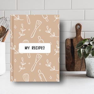 Könnte beinhalten: Ein tannenes Rezeptbuch mit einem weißen Einband, der ein sich wiederholendes Muster aus Küchenutensilien und Blättern zeigt. Der Einband trägt den Text "MY RECIPES" in einem weißen Kasten.