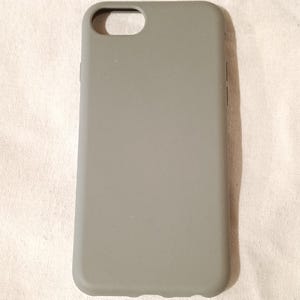 Könnte beinhalten: Eine hellgraue Handyhülle mit Aussparungen für die Kamera und andere Telefonfunktionen. Die Hülle besteht aus einem glatten Material und hat ein schlichtes, minimalistisches Design. Die Hülle soll das Telefon vor Kratzern und Beschädigungen schützen.