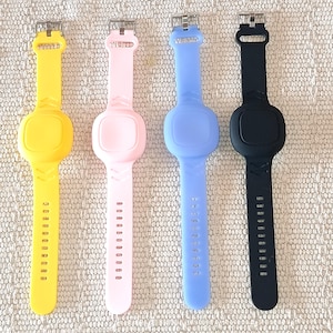 May include: Four silicone wristwatches in yellow, pink, light blue, and black. Each watch has a rectangular face and a strap with adjustment holes. The watches are displayed on a textured, neutral-toned surface.