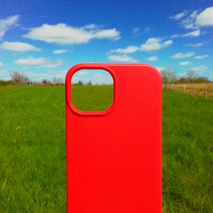 Op de afbeelding: Een felrode telefoonhoes met een uitsparing voor de camera. De hoes is voor een groen grasveld met een blauwe lucht en witte wolken op de achtergrond.