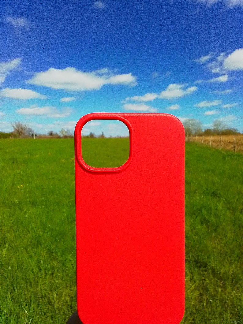 Op de afbeelding: Een felrode telefoonhoes met een uitsparing voor de camera. De hoes is voor een groen grasveld met een blauwe lucht en witte wolken op de achtergrond.