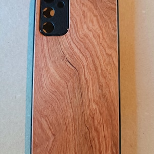 May include: A wooden-textured phone case with a black camera module. The case has a reddish-brown wood grain pattern and a black border. The phone case is rectangular and designed to protect a smartphone.