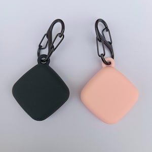 May include: Two silicone cases for wireless earbuds, one black and one pink, with black metal clips attached.