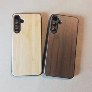 Könnte beinhalten: Zwei Handyhüllen aus Holz. Eine Hülle ist hellbraun mit einem Bambusmuster und die andere ist dunkelbraun mit einem Walnussmuster. Beide Hüllen haben einen schwarzen Rand um die Kanten und einen Ausschnitt für die Kamera.