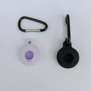 Op de afbeelding: Een set van drie items: een zwarte karabijnhaak, een lichtpaarse ronde hoes met een paars midden en een zwarte ronde hoes met een karabijnhaak. De hoesjes hebben onderaan drie kleine gaatjes.
