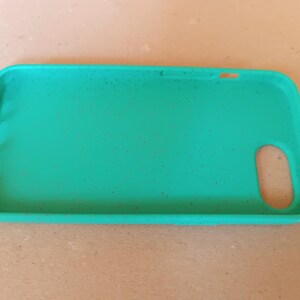 May include: A teal phone case with a rectangular shape and rounded corners. The case has cutouts for the camera, buttons, and charging port. Small dark specks are scattered across the surface. The case is made of a flexible material.