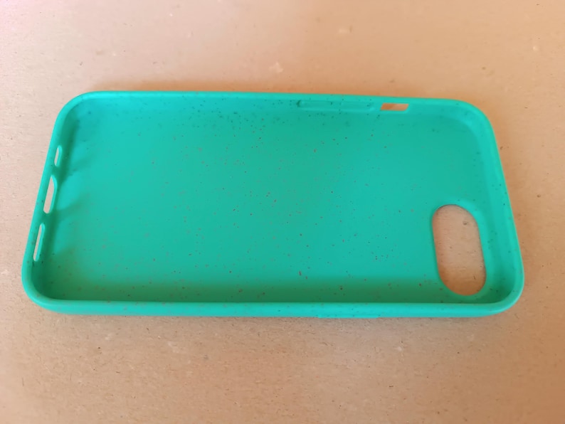 May include: A teal phone case with a rectangular shape and rounded corners. The case has cutouts for the camera, buttons, and charging port. Small dark specks are scattered across the surface. The case is made of a flexible material.