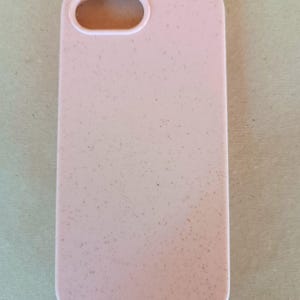 May include: A light pink phone case with a speckled texture. The case has a rectangular shape with rounded corners and an oval cutout for the camera. The case is designed to protect a smartphone.