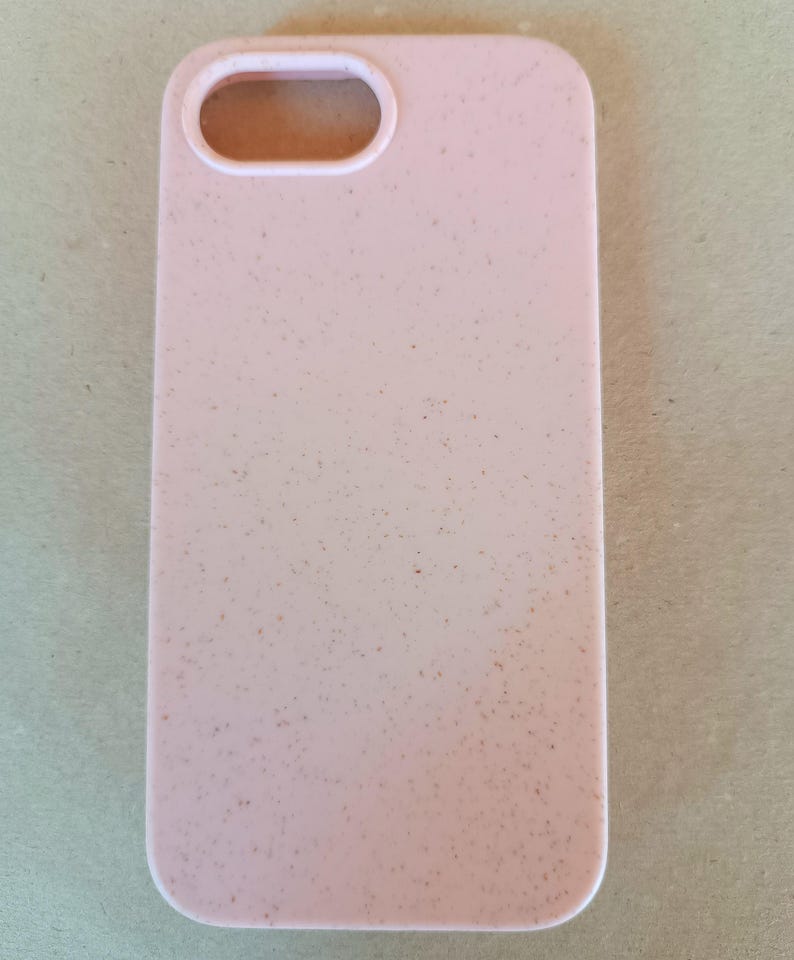 May include: A light pink phone case with a speckled texture. The case has a rectangular shape with rounded corners and an oval cutout for the camera. The case is designed to protect a smartphone.