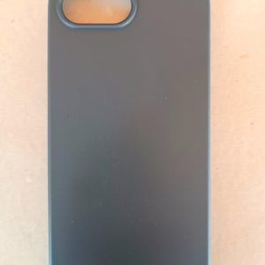 May include: A matte black phone case with a rectangular shape and rounded corners. The case has an oval opening for the camera lens. The overall design is minimalist and sleek, offering a protective cover for a smartphone.