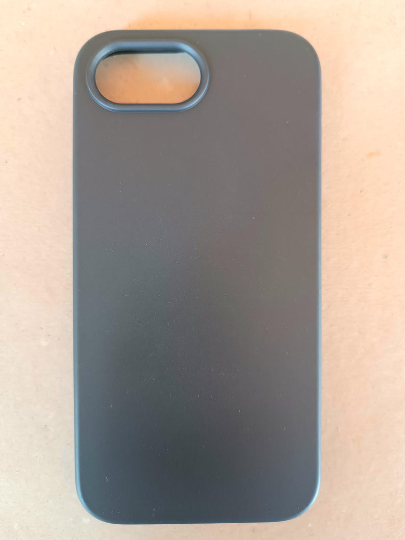 May include: A matte black phone case with a rectangular shape and rounded corners. The case has an oval opening for the camera lens. The overall design is minimalist and sleek, offering a protective cover for a smartphone.
