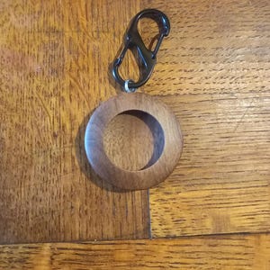 May include: A wooden keyring with a circular design and a black metal clasp. The keyring is made of light brown wood and has a smooth finish. The clasp is attached to the keyring with a small metal ring.