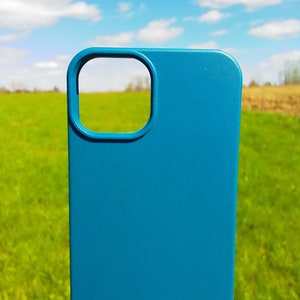 Op de afbeelding: Een blauwe telefoonhoes met een uitsparing voor de camera. De hoes is voor een groen grasveld met een blauwe lucht en witte wolken op de achtergrond.