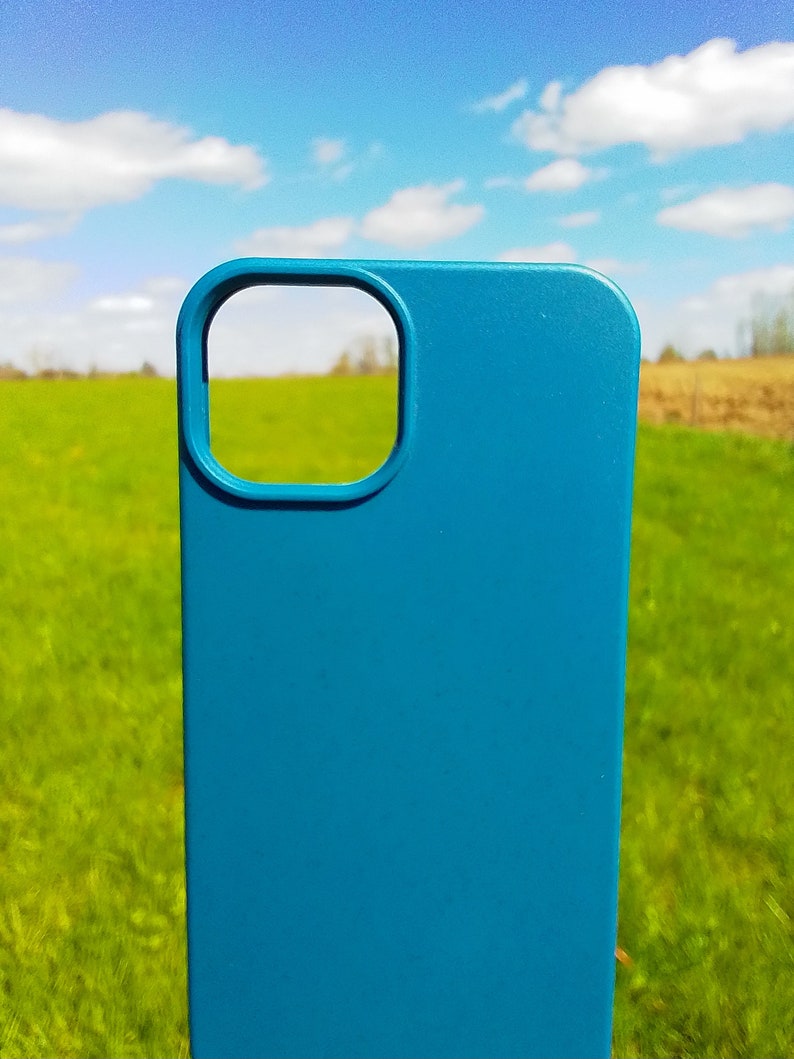 Op de afbeelding: Een blauwe telefoonhoes met een uitsparing voor de camera. De hoes is voor een groen grasveld met een blauwe lucht en witte wolken op de achtergrond.