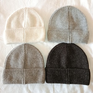 Peut inclure: Quatre bonnets en tricot côtelé crème, gris et marron foncé sont disposés sur une surface blanche. Les bonnets ont un bord replié et une conception ajustée. Les chapeaux sont faits d'une matière douce et chaude, parfaite pour le froid.