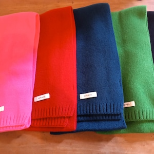 Peut inclure: Cinq écharpes en tricot de couleurs différentes, rose, rouge, bleu, vert et noir. Chaque écharpe porte une étiquette blanche avec le mot "RiverWool" imprimé dessus.