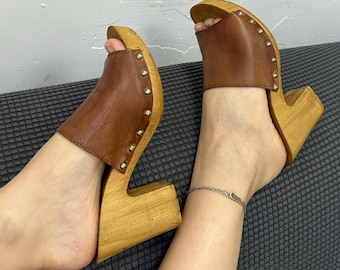 May include: Brown leather and wood clogs with open toes. The clogs have a wooden base and a brown leather upper, secured with small gold-colored studs. The heels are approximately 4 inches high.