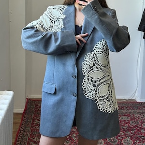 May include: Gray blazer with a cream-colored lace appliqué on the shoulder and front. The blazer has a pinstripe pattern, a notched lapel, and three buttons. The blazer is knee-length.