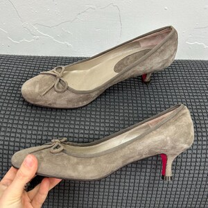 May include: A pair of taupe suede pumps with small bows on the toes. The heels are a low kitten heel with a red sole. The shoes are displayed on a dark gray textured surface.