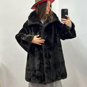 Puede incluir: Un abrigo de piel sintética marrón oscuro con cierre de botones. El abrigo tiene un cuello ancho y mangas largas. Un sombrero verde con borde rojo se lleva en la cabeza.
