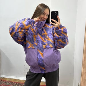 Puede incluir: Chaqueta vintage con estampado morado y naranja. La chaqueta presenta un cuello alto, una cremallera frontal y una cintura ceñida. La parte superior tiene un diseño vibrante en naranja y morado, mientras que la sección inferior es de color morado liso.