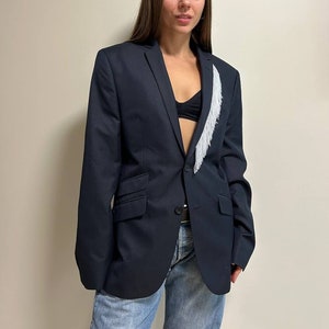 May include: A navy blue blazer with a white fringe detail on the lapel. The blazer has a single button closure and is worn over a black bra and blue jeans.
