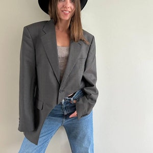 Puede incluir: Una chaqueta gris con un solo botón de cierre, puesta sobre un top beige sin mangas y unos vaqueros azules. La chaqueta tiene un dobladillo largo y asimétrico y se combina con un sombrero fedora negro.
