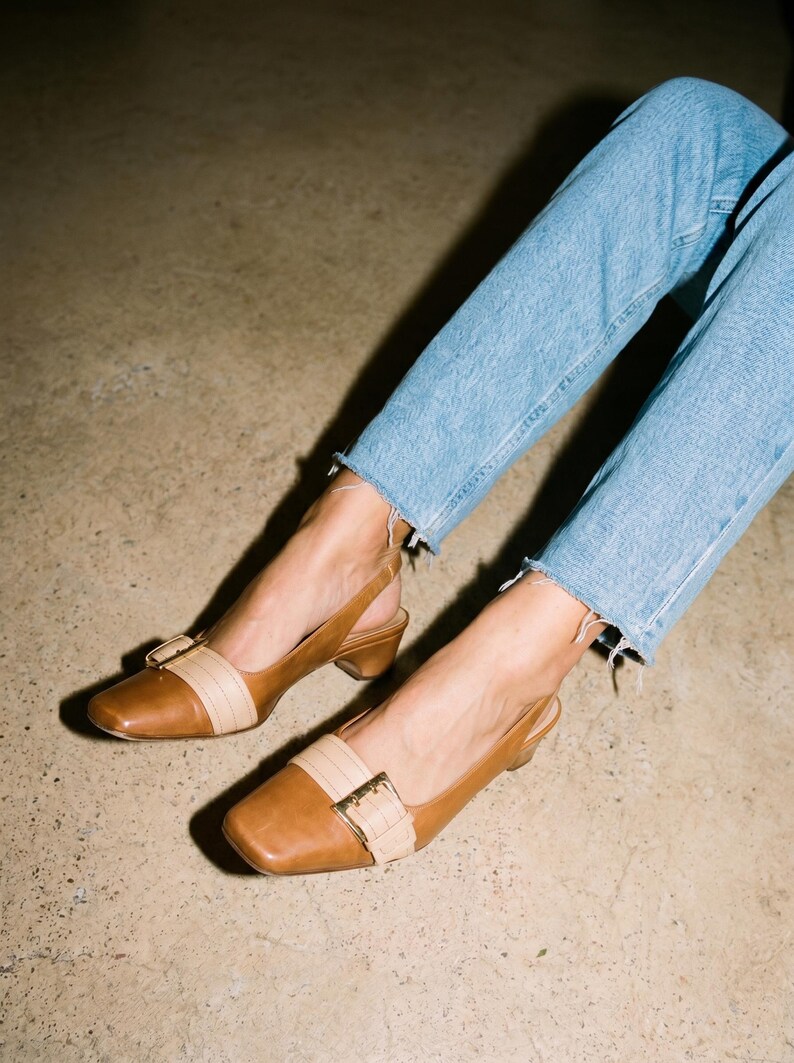 K&ouml;nnte beinhalten: Braune Slingback-Heels aus Leder mit eckiger Spitze und beigem Riemen. Die Schuhe haben einen kleinen Absatz und eine goldfarbene Schnalle. Die Person tr&auml;gt eine blaue Jeans. Die Abs&auml;tze sind ca. 5 cm hoch.