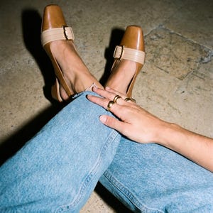 K&ouml;nnte beinhalten: Braune Slingback-Heels aus Leder mit eckiger Spitze und beigem Riemen, der mit einer goldenen Schnalle befestigt ist. Getragen mit hellblauen Jeans. Mehrere goldene Ringe schm&uuml;cken die Finger.