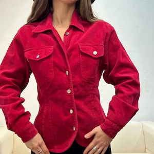 Puede incluir: Una camisa de pana roja con botones, mangas largas y dos bolsillos de solapa. La camisa tiene una cintura ajustada y un dobladillo ligeramente curvo.