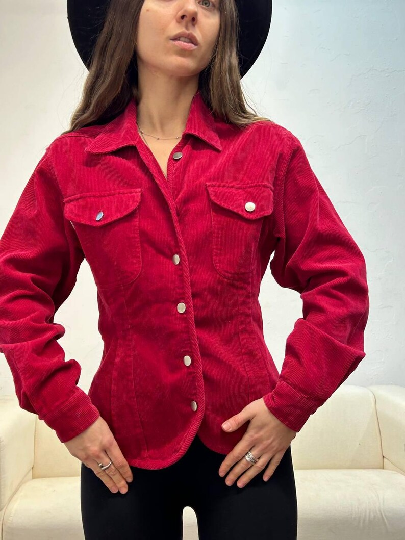 Puede incluir: Una camisa de pana roja con botones, mangas largas y dos bolsillos de solapa. La camisa tiene una cintura ajustada y un dobladillo ligeramente curvo.