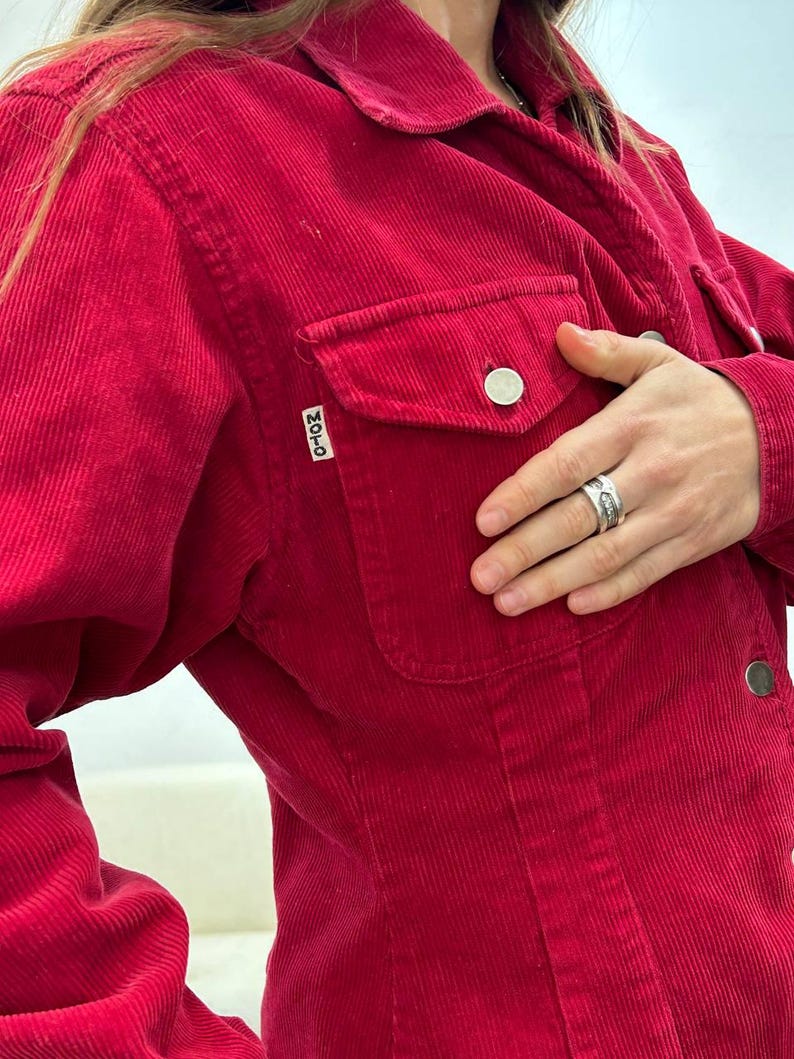 Puede incluir: Una chaqueta de pana roja con cierre de botones y un solo bolsillo en el pecho izquierdo. El bolsillo tiene una peque&ntilde;a etiqueta "MOTO".