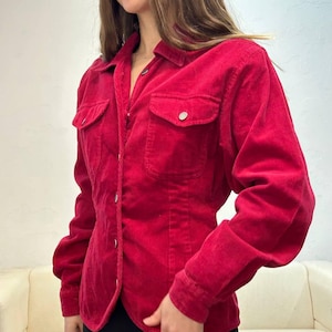 Puede incluir: Una camisa de pana roja con botones, mangas largas y silueta ajustada. La camisa tiene dos bolsillos de solapa en el pecho y un cierre de botones en la parte delantera.