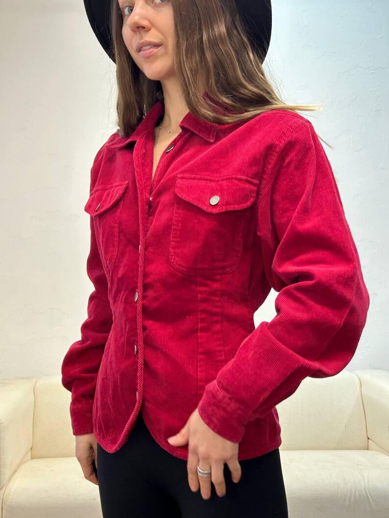 Puede incluir: Una camisa de pana roja con botones, mangas largas y silueta ajustada. La camisa tiene dos bolsillos de solapa en el pecho y un cierre de botones en la parte delantera.