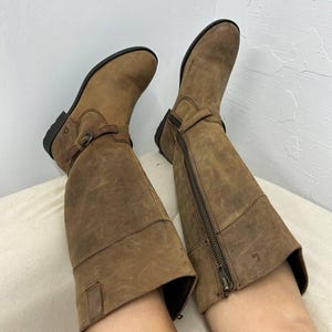 以下が含まれることがあります： サイドにジッパー、フロントにバックルストラップが付いた、ブラウンの革製のニーハイブーツのペアです。