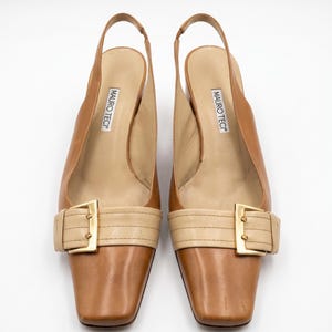 K&ouml;nnte beinhalten: Ein Paar braune Slingback-Heels aus Leder mit quadratischen Zehen. Die Schuhe haben einen beigefarbenen Riemen &uuml;ber dem Fu&szlig; mit einer goldfarbenen Schnalle. Der Markenname "MAURO TECI" ist auf der Innensohle aufgedruckt.
