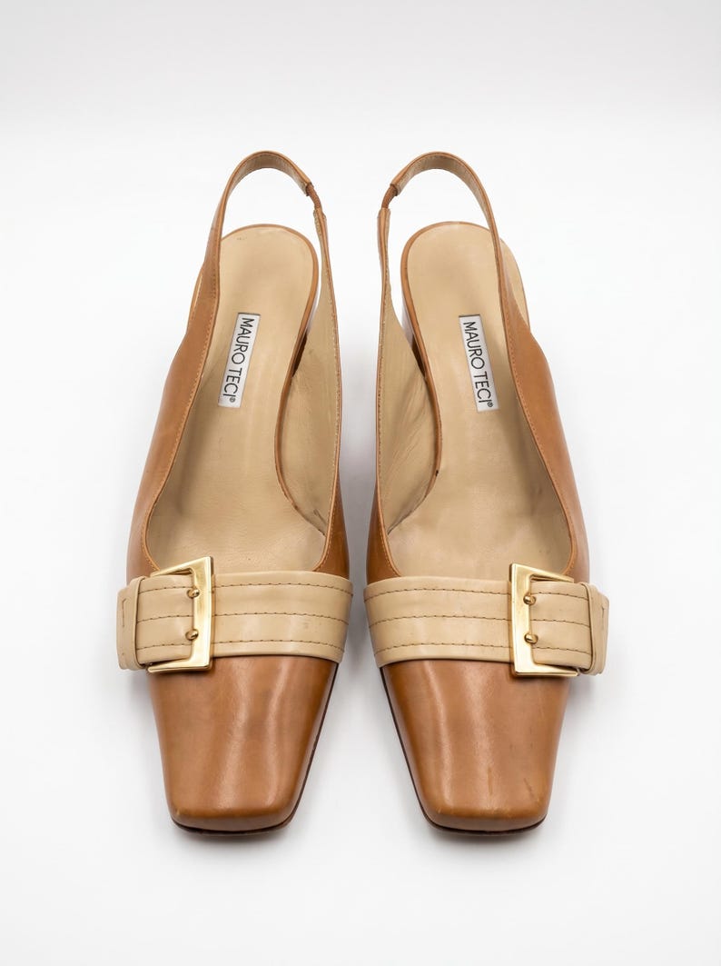 K&ouml;nnte beinhalten: Ein Paar braune Slingback-Heels aus Leder mit quadratischen Zehen. Die Schuhe haben einen beigefarbenen Riemen &uuml;ber dem Fu&szlig; mit einer goldfarbenen Schnalle. Der Markenname "MAURO TECI" ist auf der Innensohle aufgedruckt.