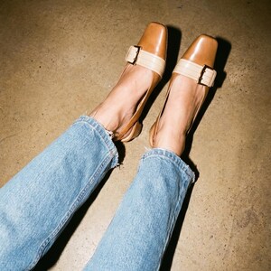 K&ouml;nnte beinhalten: Braune Lederschuhe mit eckiger Zehenpartie und beigem Riemen, der mit einer goldenen Schnalle befestigt ist. Die Schuhe werden mit hellblauen Jeans getragen.
