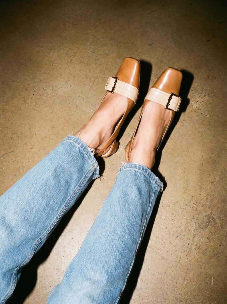 K&ouml;nnte beinhalten: Braune Lederschuhe mit eckiger Zehenpartie und beigem Riemen, der mit einer goldenen Schnalle befestigt ist. Die Schuhe werden mit hellblauen Jeans getragen.