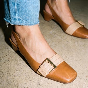 K&ouml;nnte beinhalten: Braune Leder-Slingback-Heels mit eckiger Zehenpartie und goldener Schnalle. Die Schuhe haben einen kontrastierenden beigefarbenen Riemen. Die Abs&auml;tze sind niedrig und blockartig. Getragen mit blauen Jeans.